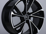 A glossy black alloy wheel with silver accents stands vertically, featuring a central emblem with four interlocked rings, set within a plain studio background.