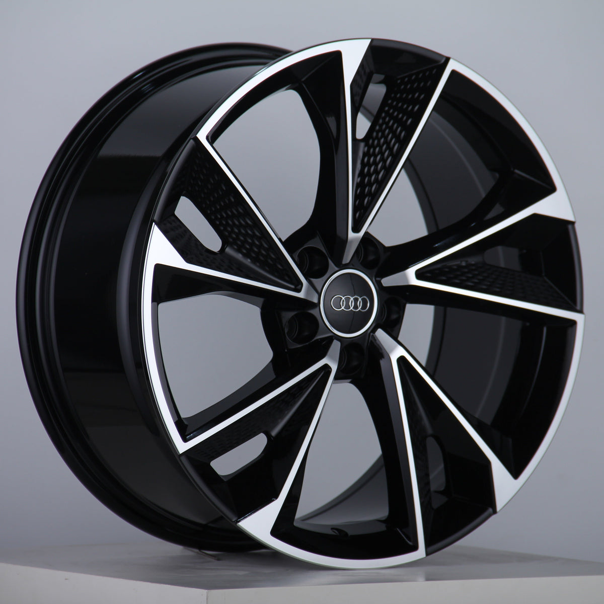 A black and silver alloy car wheel with multiple angled spokes, featuring an Audi logo at the center, is displayed against a plain gray background.