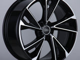A black and silver alloy wheel with a five-spoke design features the Audi logo at the center, displayed on a simple white platform against a plain gray background.