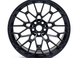 A black alloy wheel with a complex mesh design sits against a white background. The center features a small logo with blue and red stripes.