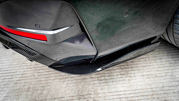 Car bumper features a red reflector and carbon fiber-like trim, positioned near a rear tire. The vehicle is parked on a grooved concrete surface.