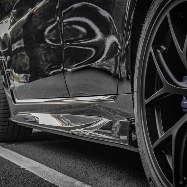 A black car's side view, showing glossy bodywork and a detailed wheel with a BMW logo, parked on asphalt near trees. Visible text includes "SOCODA."