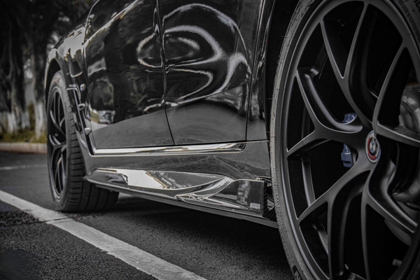A black car's side view, showing glossy bodywork and a detailed wheel with a BMW logo, parked on asphalt near trees. Visible text includes "SOCODA."