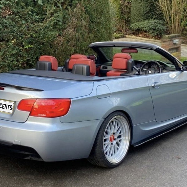 Convertible BMW with red interior parked on a driveway, surrounded by lush hedges and trees. The rear license plate reads "CARBON ACCENTS."