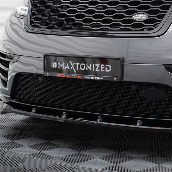 A car's front bumper with a grille displays a plaque reading "#MAXTONIZED Porsche Centrum Poznań" in a patterned indoor setting.