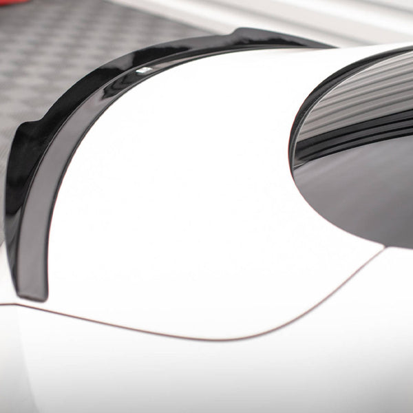 A sleek black spoiler rests on the rear of a white car, positioned in an indoor space with a checkered floor, adjacent to red car components.