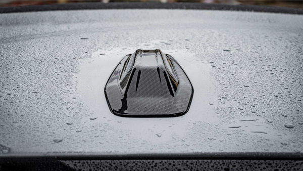 A black, shark fin car antenna rests on a gray, wet car roof, with raindrops scattered across the surface creating a textured appearance.