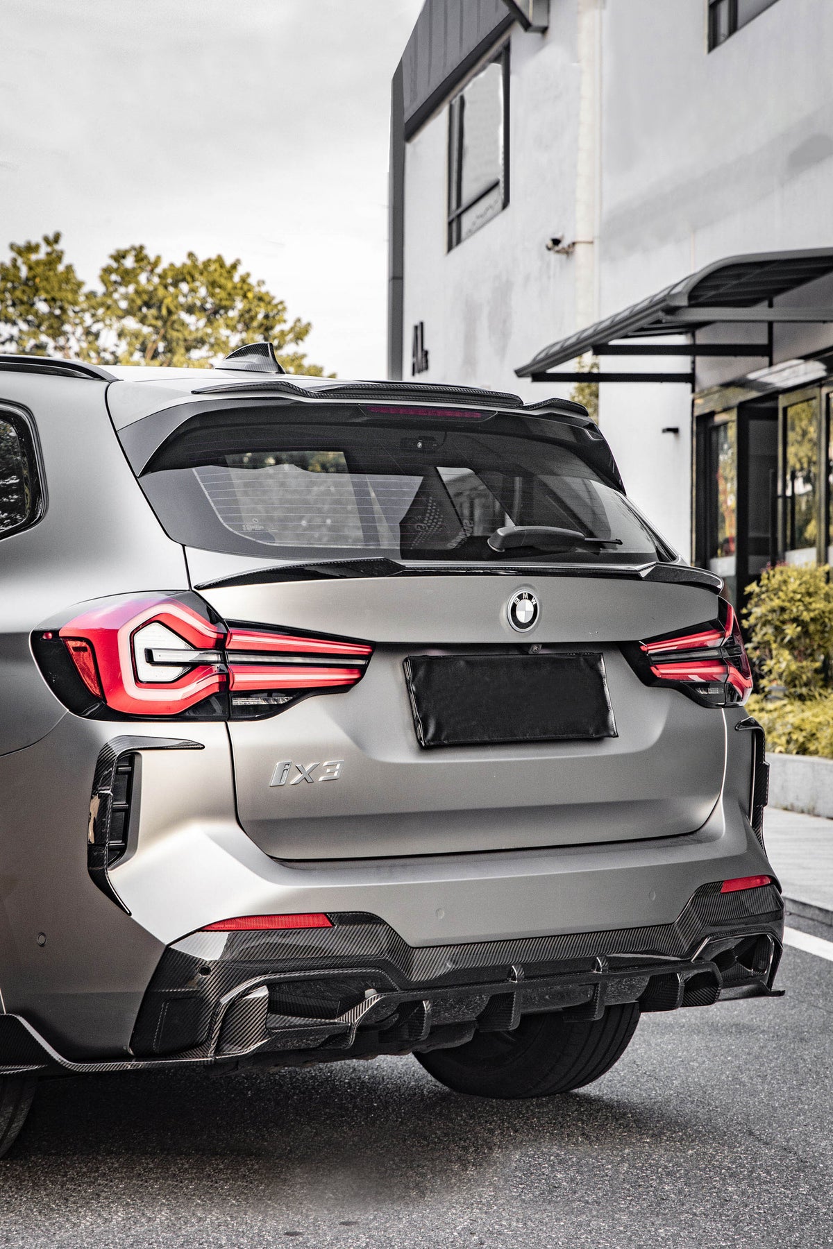 A BMW iX3 parked on a street, featuring prominent taillights and a sleek rear design. It's positioned near a modern white building with green foliage in the background.