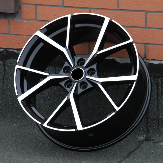 A shiny black alloy wheel rests against a brick wall. It features a modern, angular spoke design with silver accents, highlighting a sleek and stylish appearance.