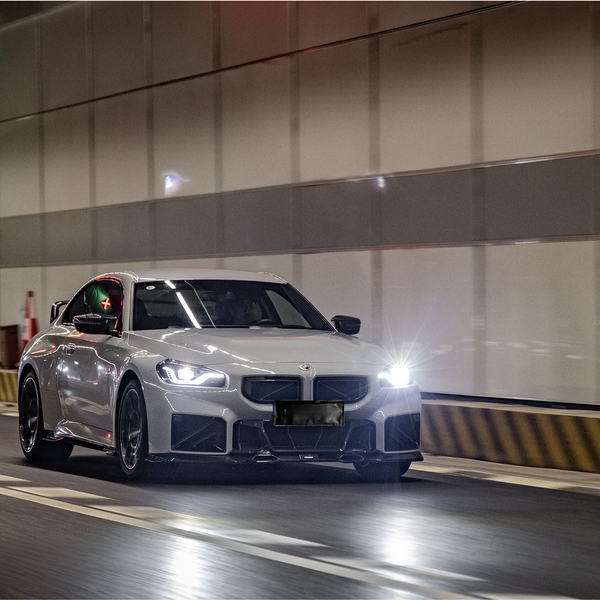 A white sports car speeds through a well-lit tunnel, headlights illuminating the path. The tunnel's striped walls and traffic cones provide a sense of motion and urban environment.