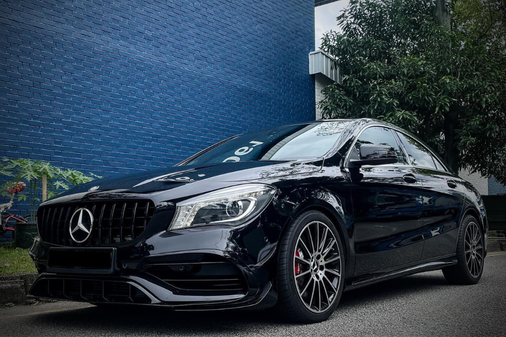 A black Mercedes-Benz car is parked on a street beside a blue brick wall, with greenery nearby and a tree overhead, conveying a sleek, urban environment.