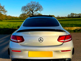 A silver Mercedes-Benz car is parked on a road, facing away. The context is a scenic countryside with green fields and a leafless tree under a clear blue sky.