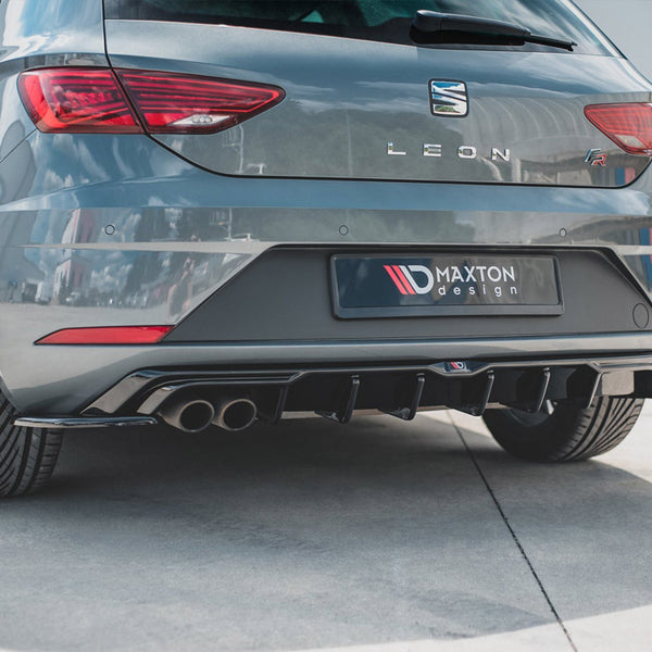 A gray hatchback car, parked on a smooth surface, displays a sporty rear bumper with dual exhausts. The license plate reads "MAXTON design," surrounded by a bright outdoor environment.
