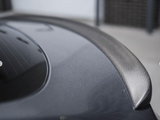 A sleek carbon fiber spoiler rests on a gray car's trunk, reflecting light subtly. The car is parked indoors against a neutral backdrop with lattice panels in the distance.