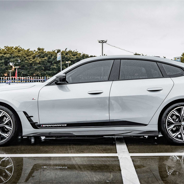 A silver sports car is parked on a wet, reflective surface with "M PERFORMANCE" branding. It's surrounded by a fenced area, trees, and a multi-story building in the background.