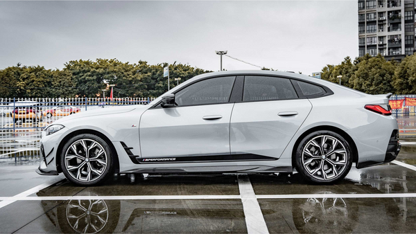 A silver sports car is parked on a wet, reflective surface with "M PERFORMANCE" branding. It's surrounded by a fenced area, trees, and a multi-story building in the background.