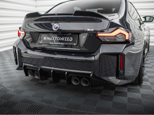 A sleek, black BMW M2 displays its rear design, featuring four exhaust pipes. It is parked on a patterned floor with white panelled walls. Text on the license plate reads "#MAXTONIZED, Dealer BMW M Cars."