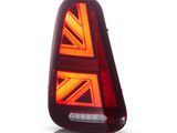 A red automotive tail light with illuminated chevron patterns is positioned upright against a plain white background. It features a horizontal white reflector at the bottom.
