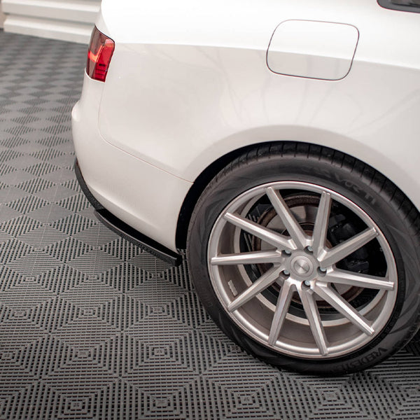 Car rear wheel is parked on a textured gray garage floor. The white vehicle's body features a visible fuel cap, with red taillight on display in the upper corner.