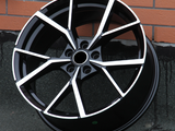 A black alloy wheel with a modern, angular design featuring silver accents rests against a brick wall on a textured concrete surface.