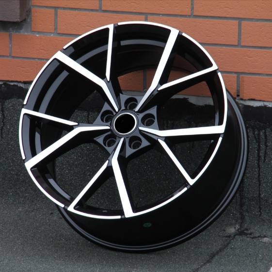 A black alloy wheel with a modern, angular design featuring silver accents rests against a brick wall on a textured concrete surface.
