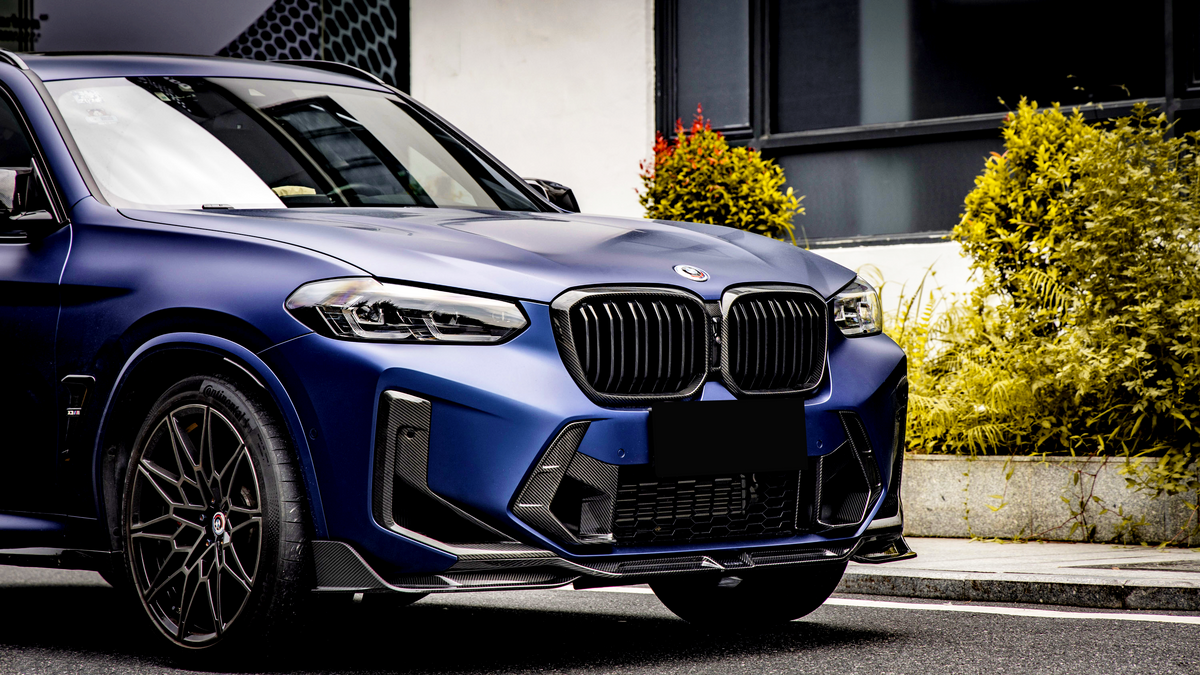 A blue SUV with a visible front grille and emblem is parked on a street. The scene includes surrounding greenery and a building with black-framed windows.