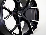 A glossy black car wheel with intricate spoke design displays an Audi logo in the center, set against a plain white background.