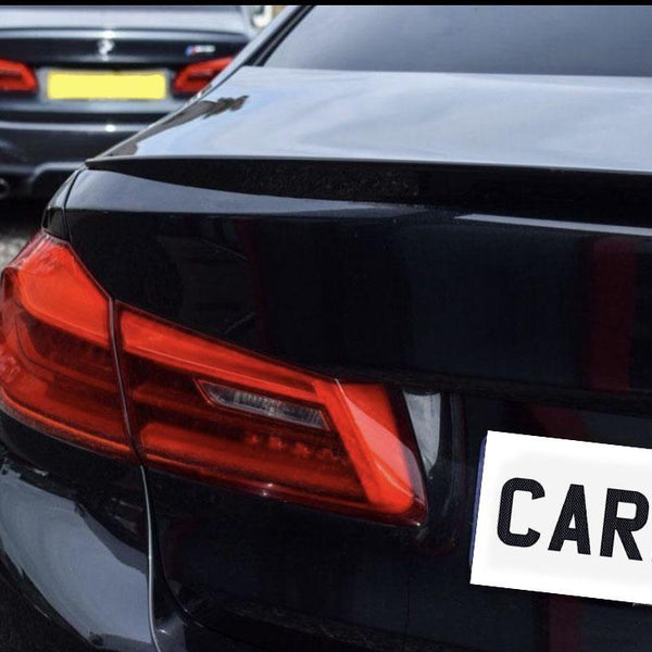 A dark car with red taillights is parked on a gravel surface, with another vehicle visible in the background. The visible text reads "CARBO."