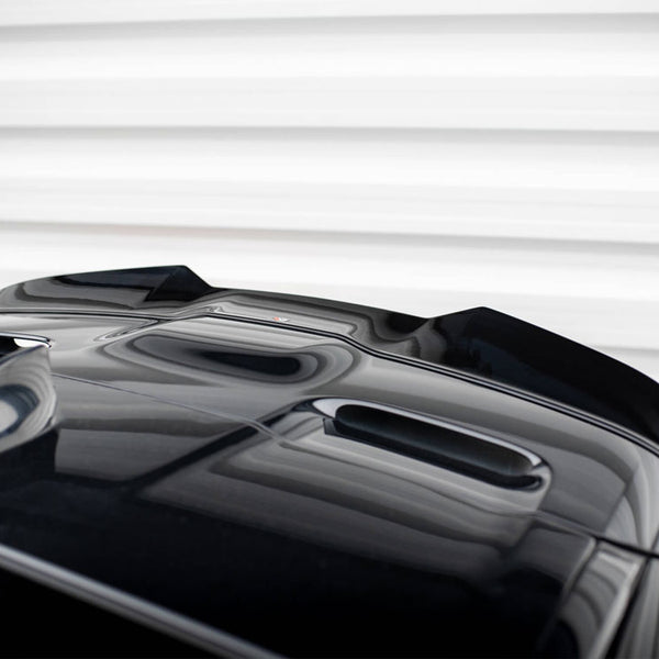 A glossy black car's hood with streamlined air vents sits stationary, reflecting light. It's positioned against a backdrop of horizontal, white, textured panels.