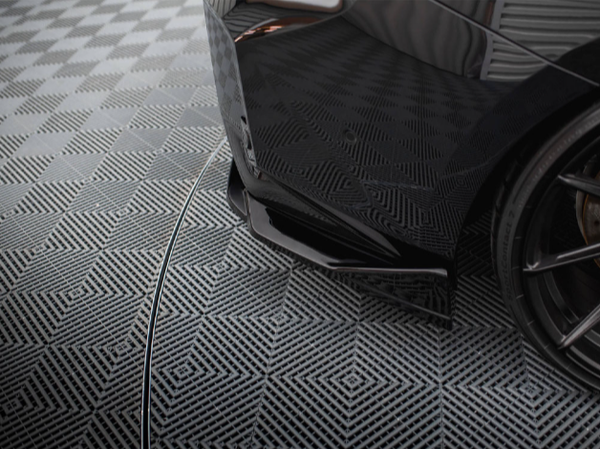 A sleek black car's rear bumper is parked on a patterned grid floor, surrounded by a symmetrical, geometric design in a well-lit, indoor setting.