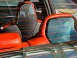 Car seats with carbon fiber backs and red leather accents, viewed through a rear window with orange defrost lines, inside a parked vehicle illuminated by sunlight.