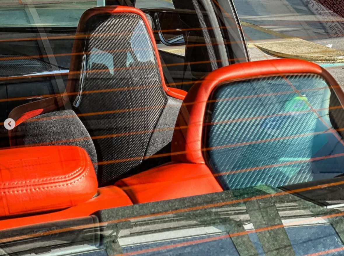 Car seats with carbon fiber backs and red leather accents, viewed through a rear window with orange defrost lines, inside a parked vehicle illuminated by sunlight.