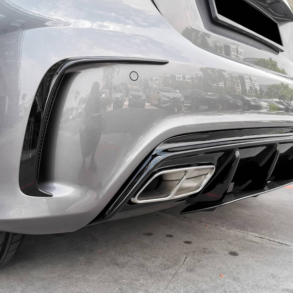 A metallic gray car's rear bumper and exhaust are shown, reflecting surrounding buildings and parked cars. The vehicle is positioned on a concrete surface under a partially cloudy sky.