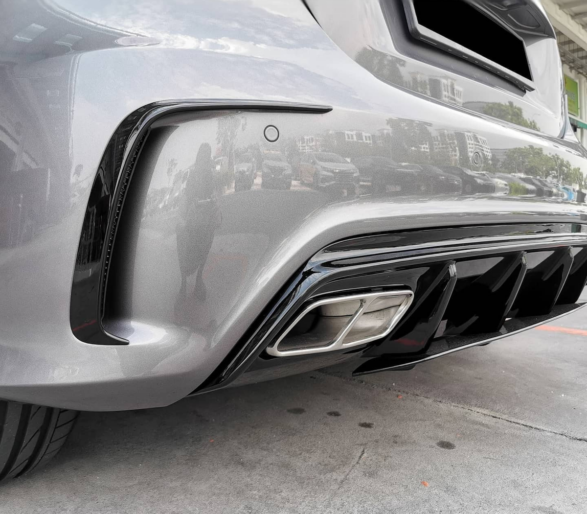 A metallic gray car's rear bumper and exhaust are shown, reflecting surrounding buildings and parked cars. The vehicle is positioned on a concrete surface under a partially cloudy sky.