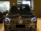 A black SUV with illuminated headlights is parked indoors, featuring a prominent front grille displaying the Mercedes-Benz logo. The showroom's ambient lighting reflects on its glossy surface.