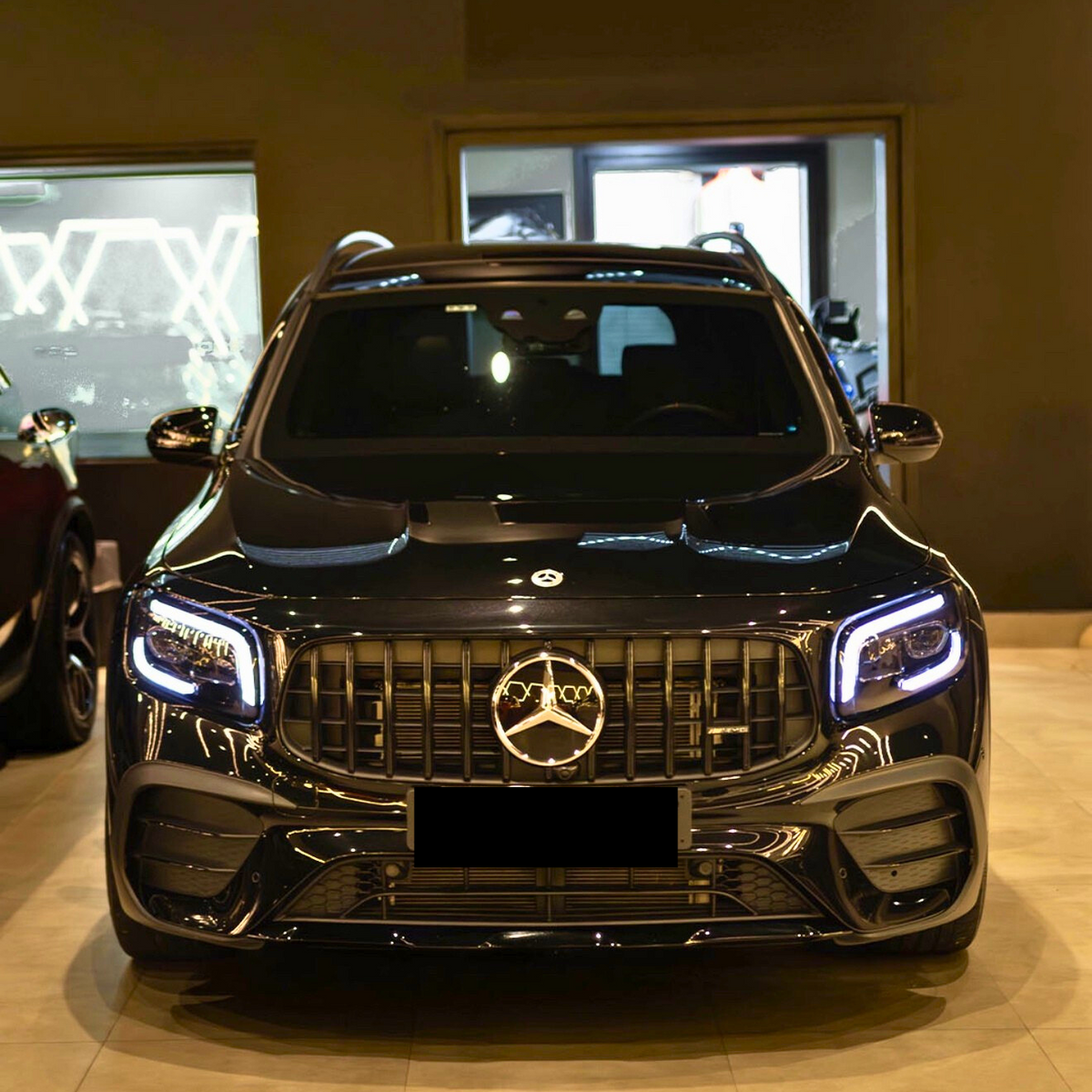 A black SUV with illuminated headlights is parked indoors, featuring a prominent front grille displaying the Mercedes-Benz logo. The showroom's ambient lighting reflects on its glossy surface.