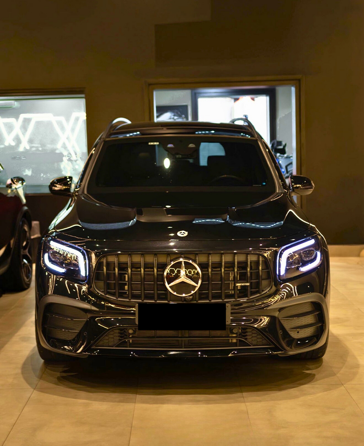 A black SUV with illuminated headlights is parked indoors, featuring a prominent front grille displaying the Mercedes-Benz logo. The showroom's ambient lighting reflects on its glossy surface.