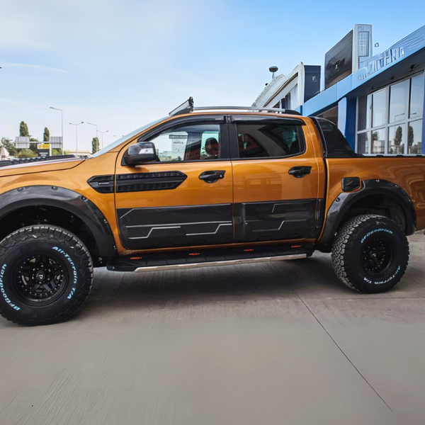 Orange pickup truck parked by a building, featuring rugged tires with "BFGoodrich All-Terrain T/A" text, against a clear sky backdrop. Notable black detailing adorns the vehicle's sides and wheel arches.