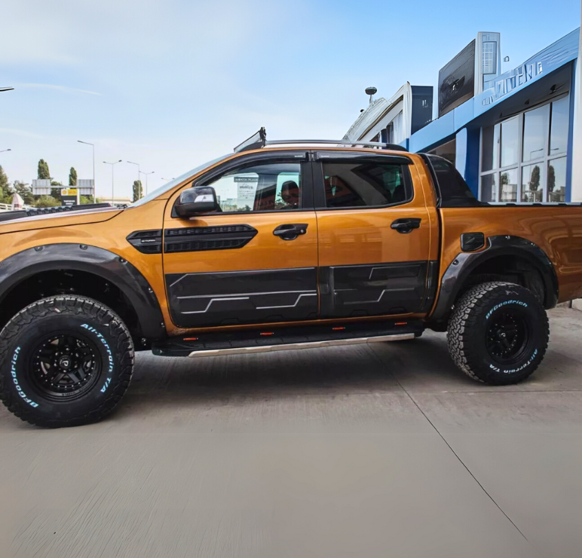 Orange pickup truck parked by a building, featuring rugged tires with "BFGoodrich All-Terrain T/A" text, against a clear sky backdrop. Notable black detailing adorns the vehicle's sides and wheel arches.