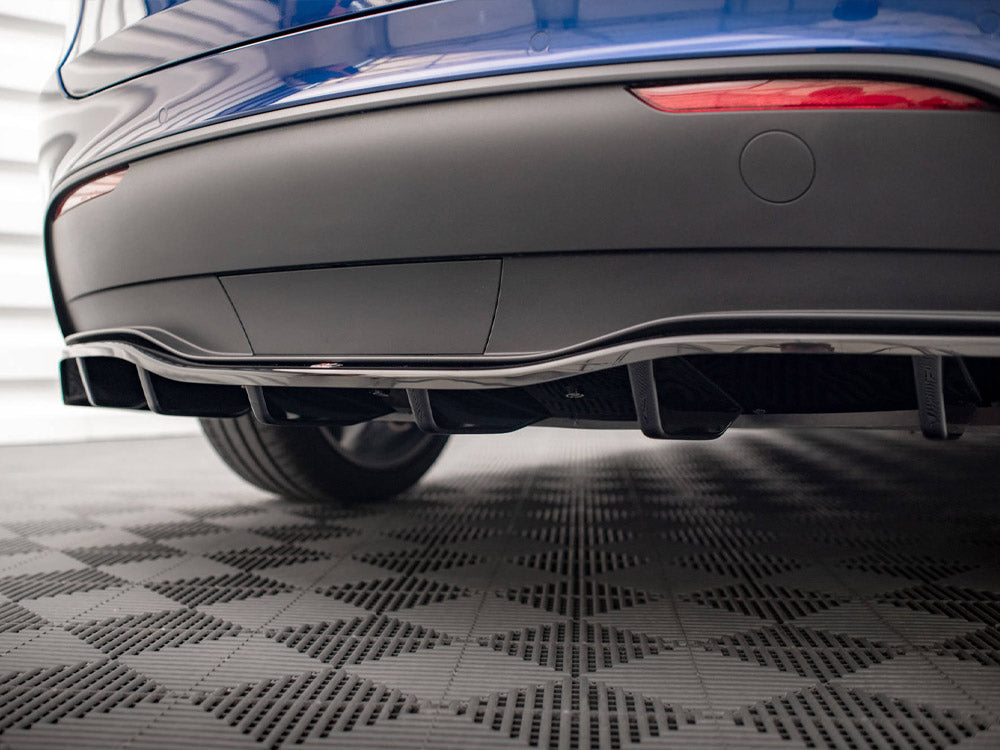A car's rear diffuser is positioned low, enhancing aerodynamics. It's part of a blue vehicle, parked on a textured, checkered floor. Two red reflector lights are visible above the diffuser.
