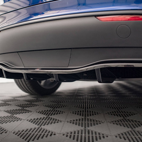 A car's rear diffuser is positioned low, enhancing aerodynamics. It's part of a blue vehicle, parked on a textured, checkered floor. Two red reflector lights are visible above the diffuser.