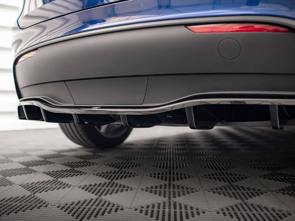 A car's rear diffuser is positioned low, enhancing aerodynamics. It's part of a blue vehicle, parked on a textured, checkered floor. Two red reflector lights are visible above the diffuser.