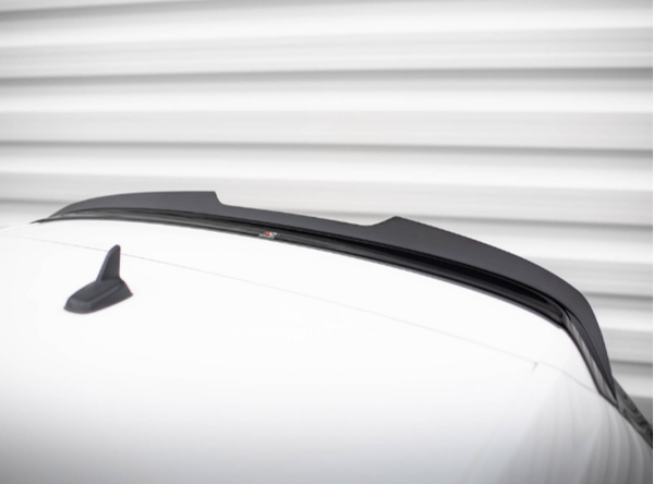 A sleek black car spoiler is mounted on a white vehicle roof, with a small shark-fin antenna nearby, set against a blurred horizontal striped background.