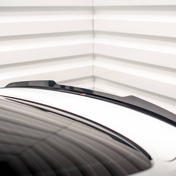 A black spoiler is positioned on a white car trunk, with a sleek design. The setting is a room with white, horizontally-lined walls, adding a modern aesthetic.