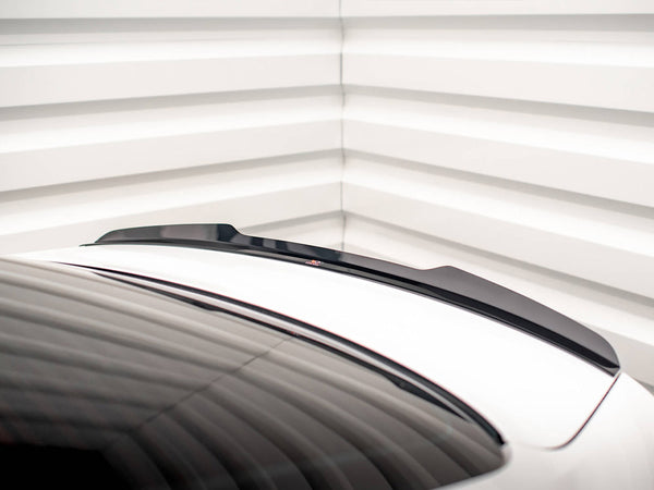 A black spoiler is positioned on a white car trunk, with a sleek design. The setting is a room with white, horizontally-lined walls, adding a modern aesthetic.