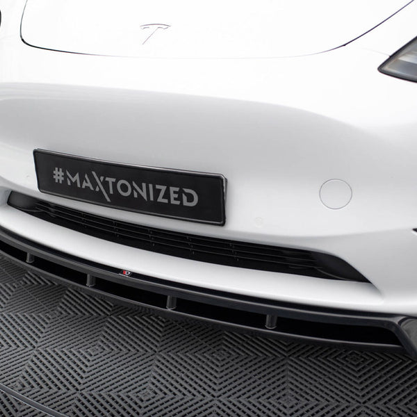 A car's front bumper with a "#Maxtonized" plate above a patterned floor, featuring sleek headlights and a prominent grille, set in a modern indoor space.