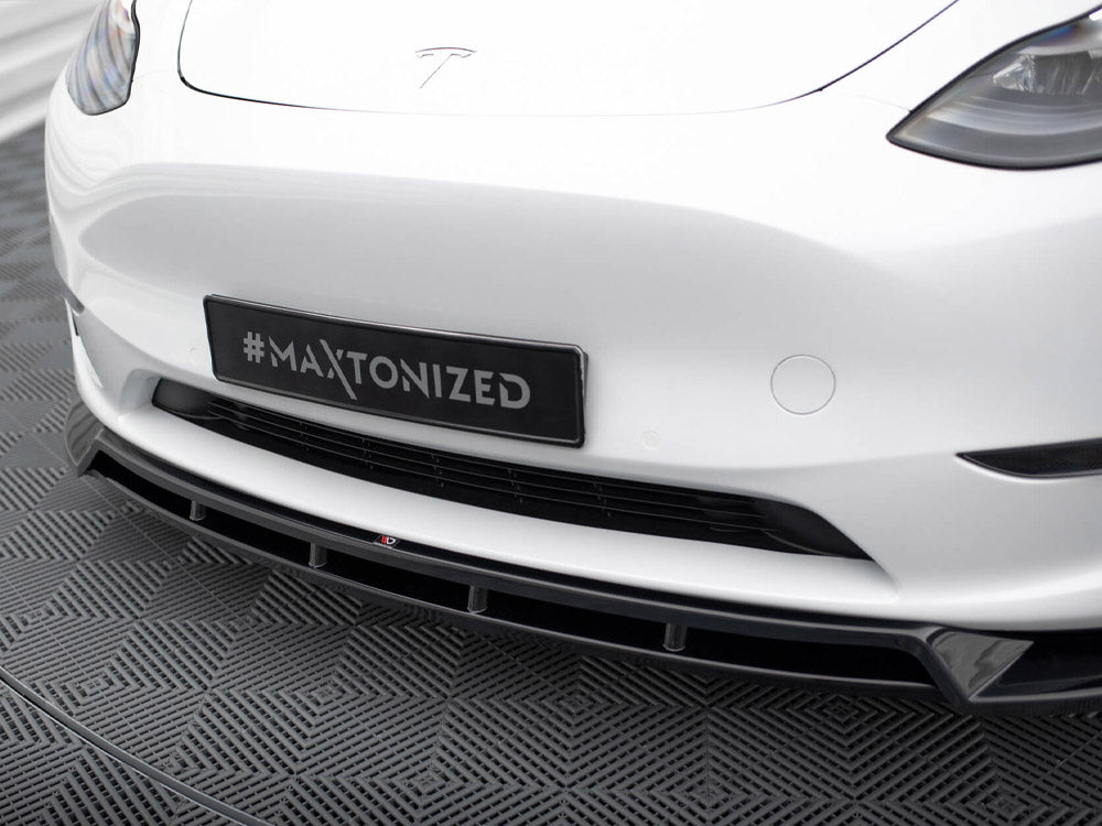 A car's front bumper with a "#Maxtonized" plate above a patterned floor, featuring sleek headlights and a prominent grille, set in a modern indoor space.
