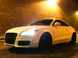 A white sports car is parked, with its headlights shining brightly, against a textured brick wall under warm streetlights, creating a dramatic nighttime urban scene.