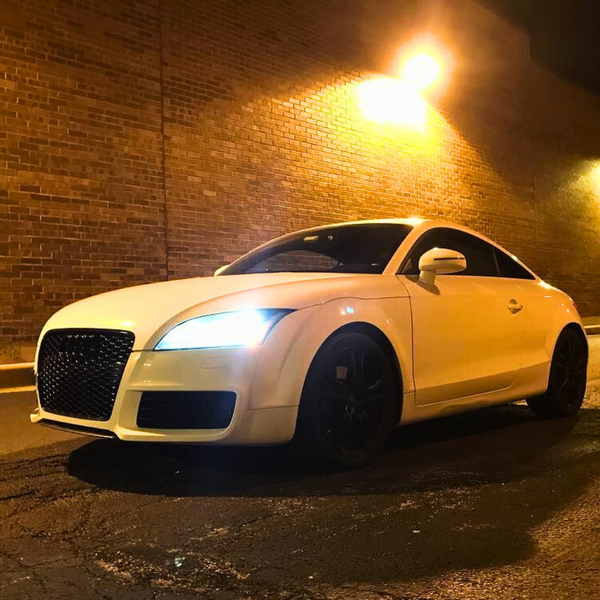 A white sports car is parked, with its headlights shining brightly, against a textured brick wall under warm streetlights, creating a dramatic nighttime urban scene.
