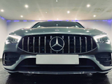 A silver car with a prominent grille and illuminated headlights is parked indoors under bright ceiling lights, creating a sleek and modern atmosphere. A Mercedes-Benz logo is centered on the grille.